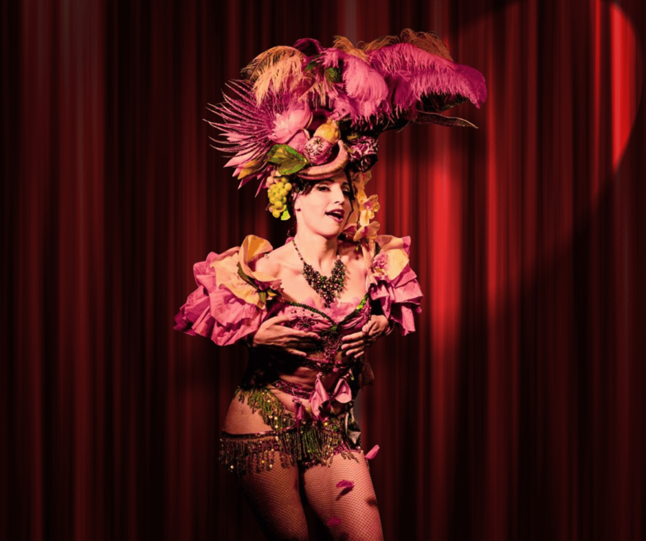 L’artista di Burlesque Candy Paradise di Cabaret Sospeso si esibisce in uno show di Burlesque. Indossa un sontuoso costume rosa con piume e copricapo scenografico sotto un riflettore caldo su sfondo di velluto rosso